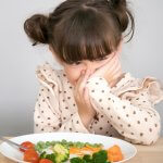 Kind sitzt vor Teller mit Gemüse und verzieht das Gesicht